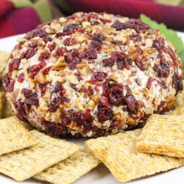 Cheese ball coated in nuts and dried cranberries, surrounded by square crackers on a white plate.