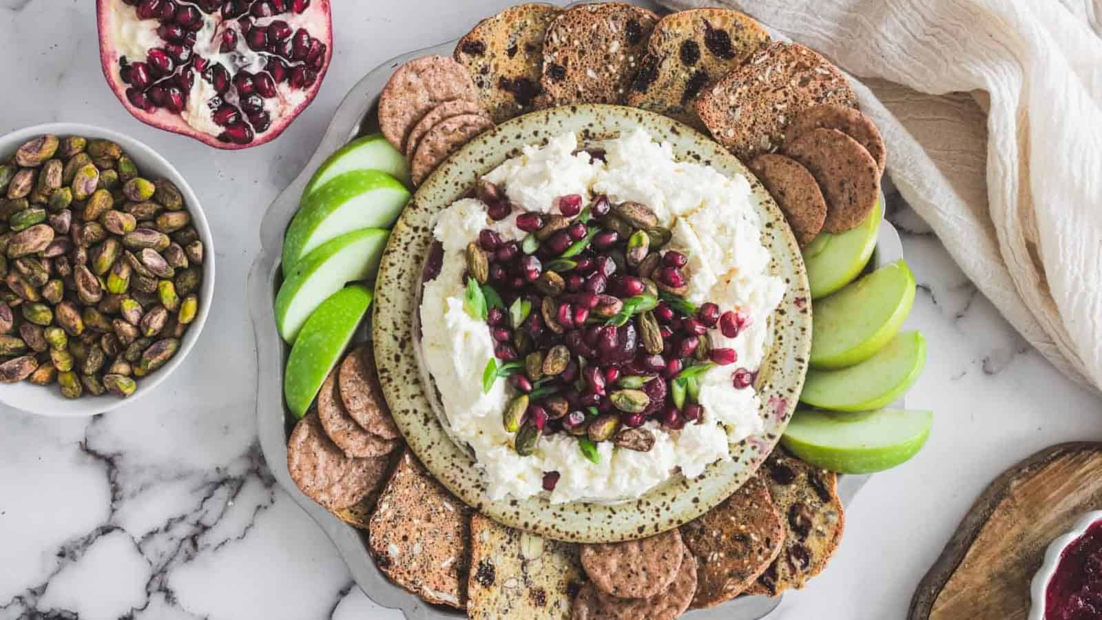 A platter with whipped cheese topped with pomegranate seeds, pistachios, apple slices, and crackers.