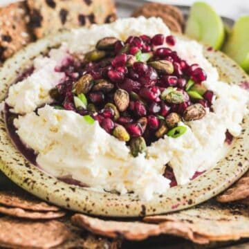 A platter with whipped cheese topped with pomegranate seeds, pistachios, apple slices, and crackers.