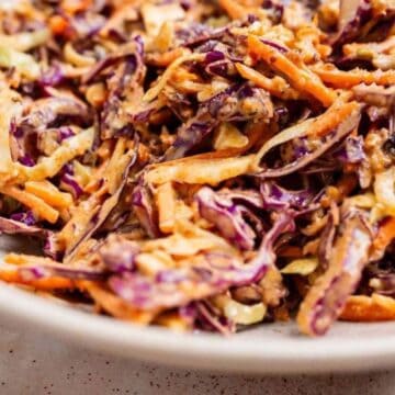 A close-up of colorful coleslaw with shredded carrots and red cabbage in a bowl with a wooden spoon.