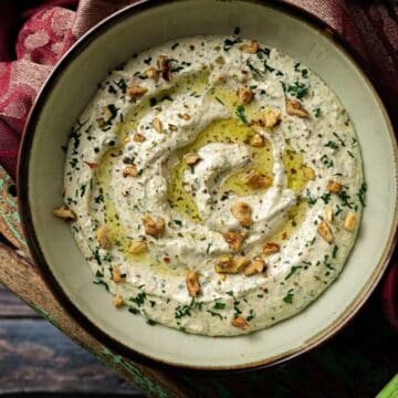 A bowl of creamy dip garnished with chopped herbs, nuts, and a drizzle of olive oil on a rustic table.