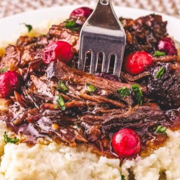 Shredded pot roast with gravy and cranberries on mashed potatoes, with a fork on a white plate.