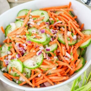 A bowl of salad with sliced cucumbers, shredded carrots, and chopped red onions on a light tablecloth.