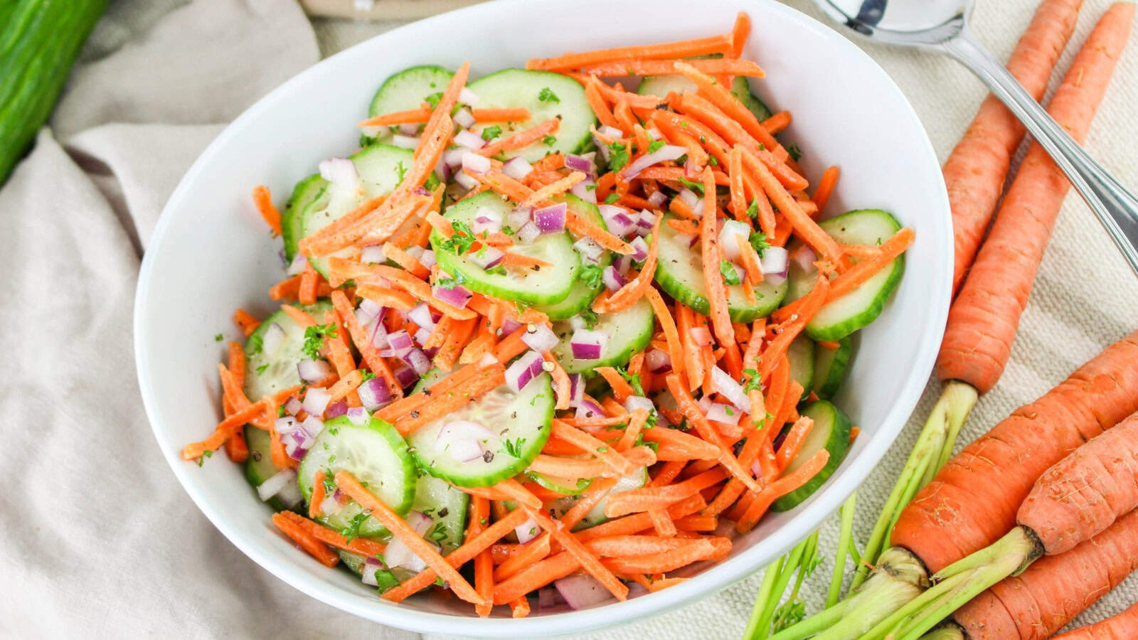 A bowl of salad with sliced cucumbers, shredded carrots, and chopped red onions on a light tablecloth.