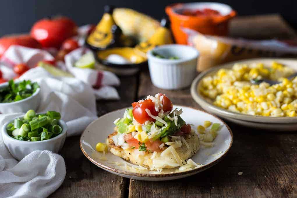 A plate with cornbread topped with cheese, tomatoes, cilantro, and vegetables, with corn and toppings nearby.
