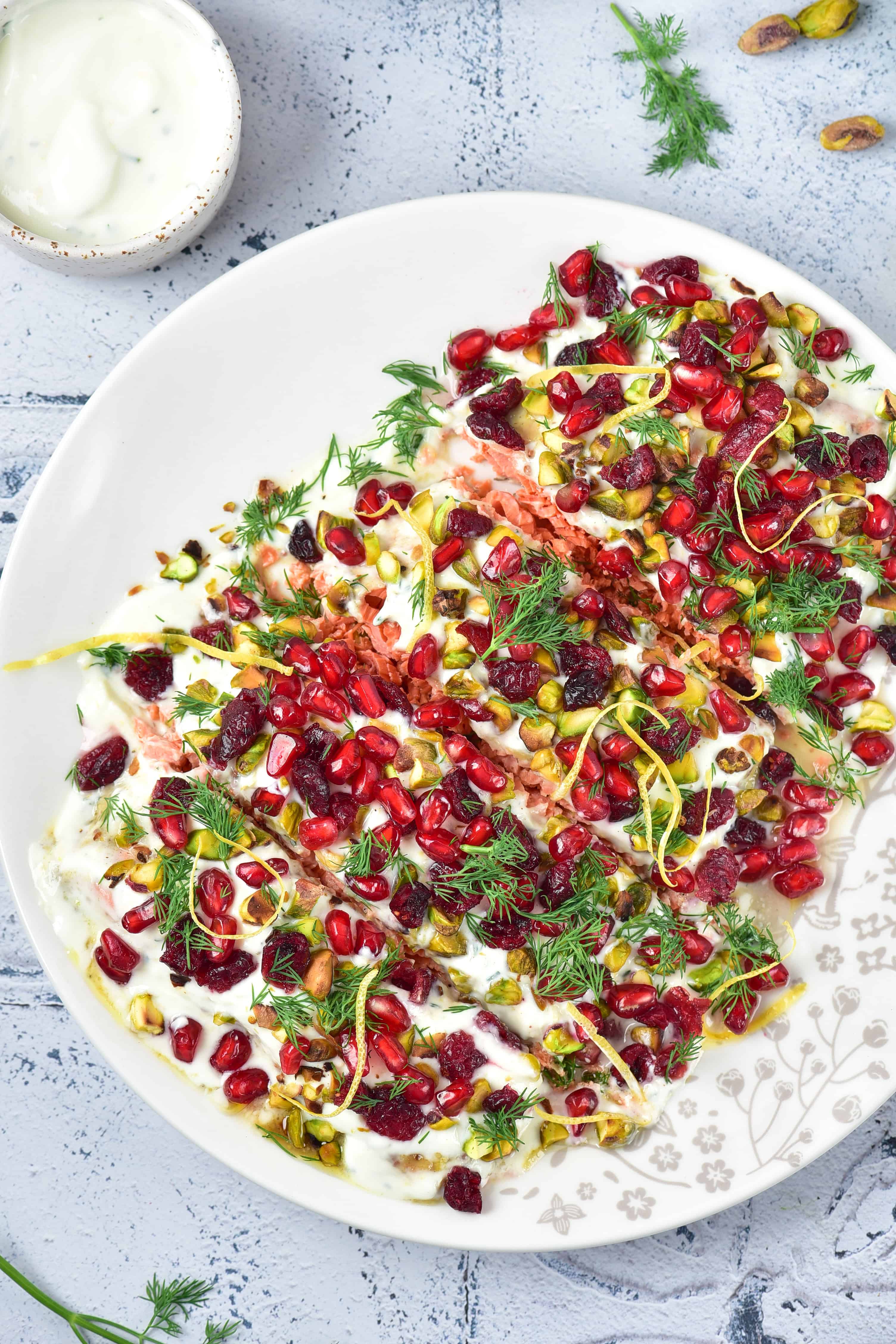 A plate of yogurt dip topped with pomegranate seeds, pistachios, dried cranberries, lemon zest, and fresh herbs.