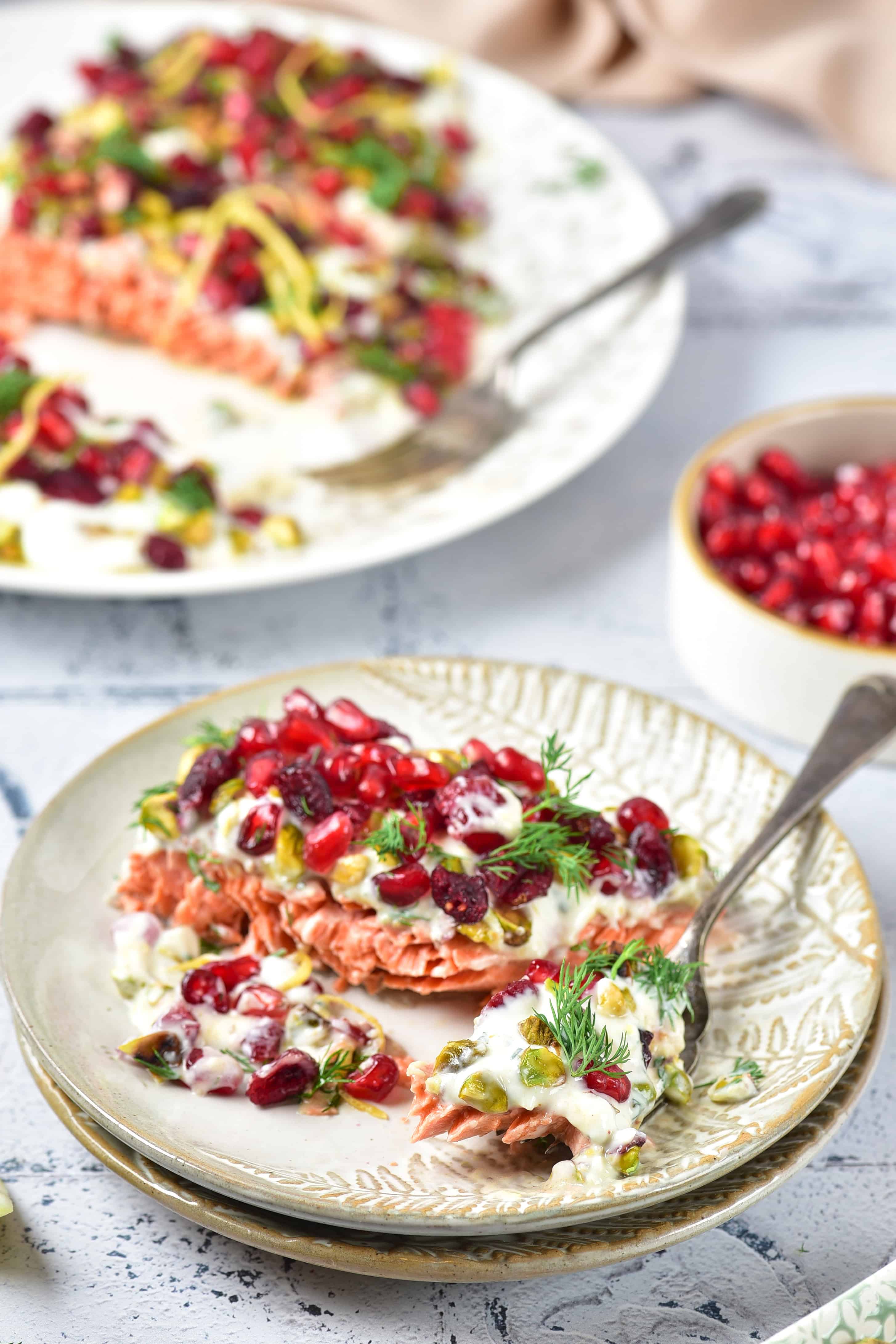 Sliced salmon topped with creamy sauce, pomegranate seeds, herbs, and lemon zest on a plate.