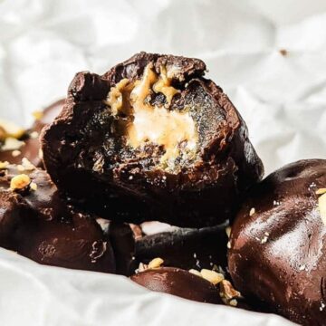 Close-up of chocolate-covered treats with creamy filling, topped with chopped nuts on white parchment paper.