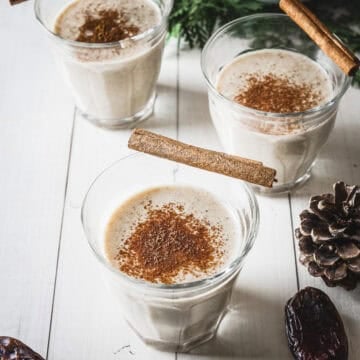 Three glasses of creamy, date sweetened eggnog topped with cinnamon sticks and a sprinkle of ground cinnamon rest elegantly on a white table.