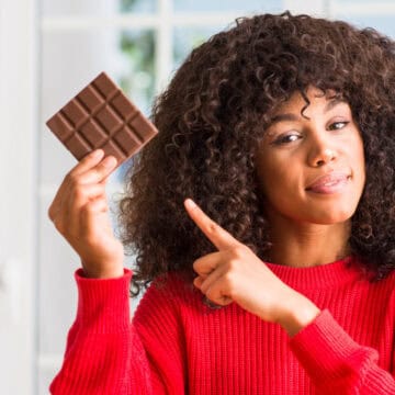 Woman in a red sweater smiling and holding up a chocolate bar while pointing at it.