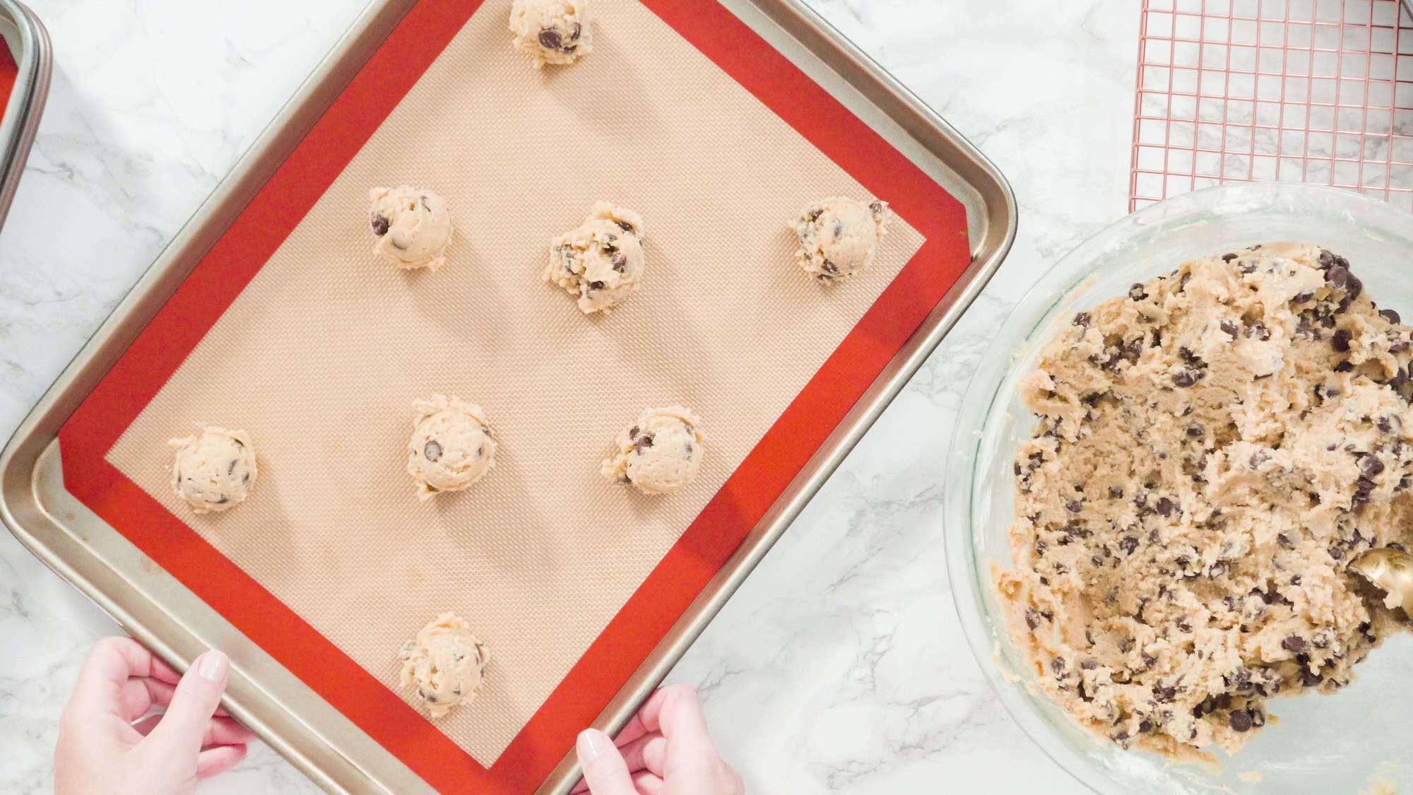 Hands holding a baking tray with scoops of chocolate chip cookie dough on a silicone mat, next to a bowl of dough—a clever pick for Gifts That Make Holiday Cleanup Easier.