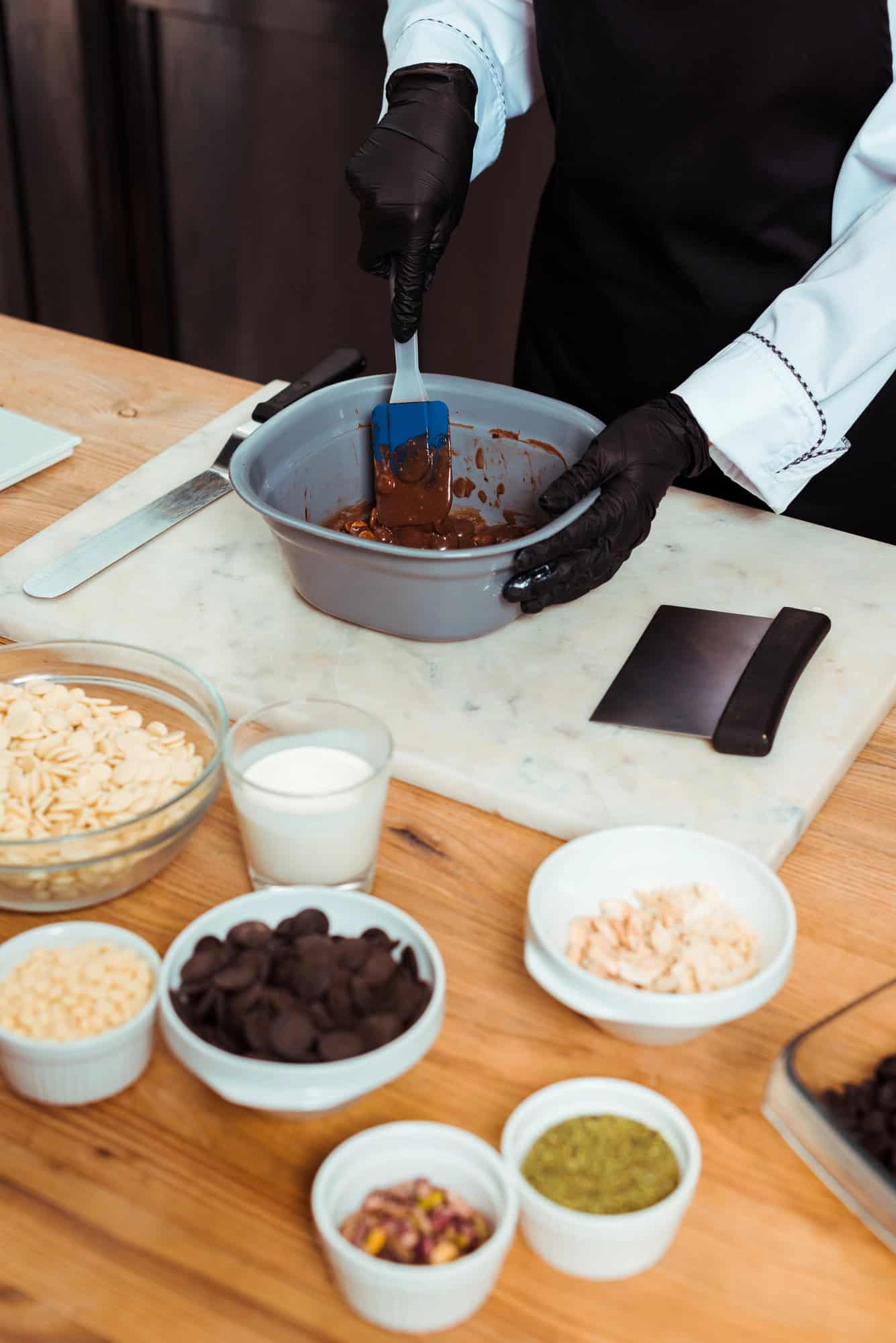Person in black gloves mixing melted chocolate in a bowl, surrounded by baking ingredients on a table—perfect inspiration for Gifts That Make Holiday Cleanup Easier.
