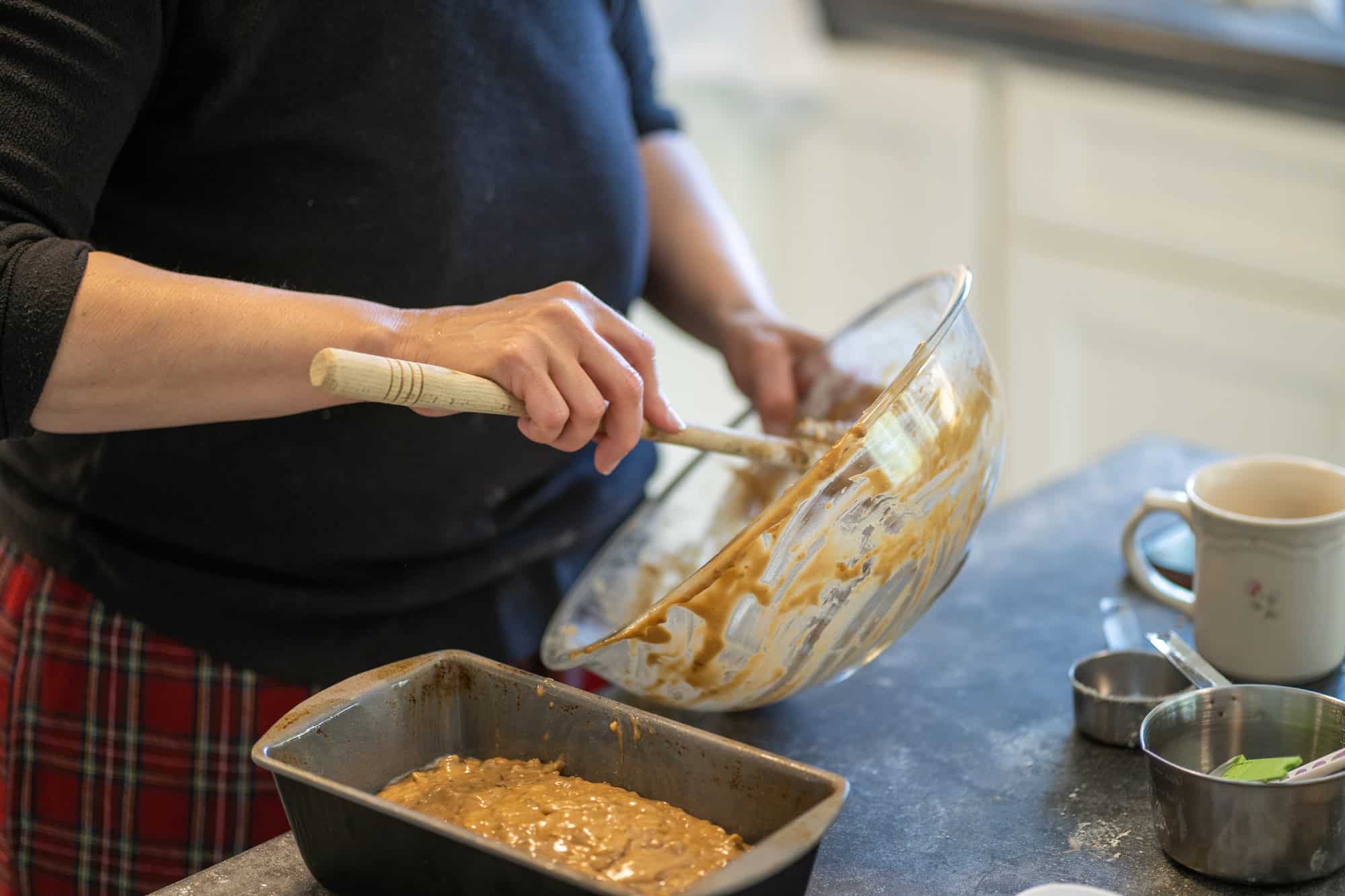 Person spooning batter from a glass bowl into a loaf pan on a kitchen counter with baking tools nearby, using Gifts That Make Holiday Cleanup Easier.