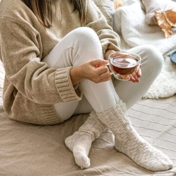 Woman in cozy socks and sweater sits on a bed holding a cup of tea, with a book and candle nearby.
