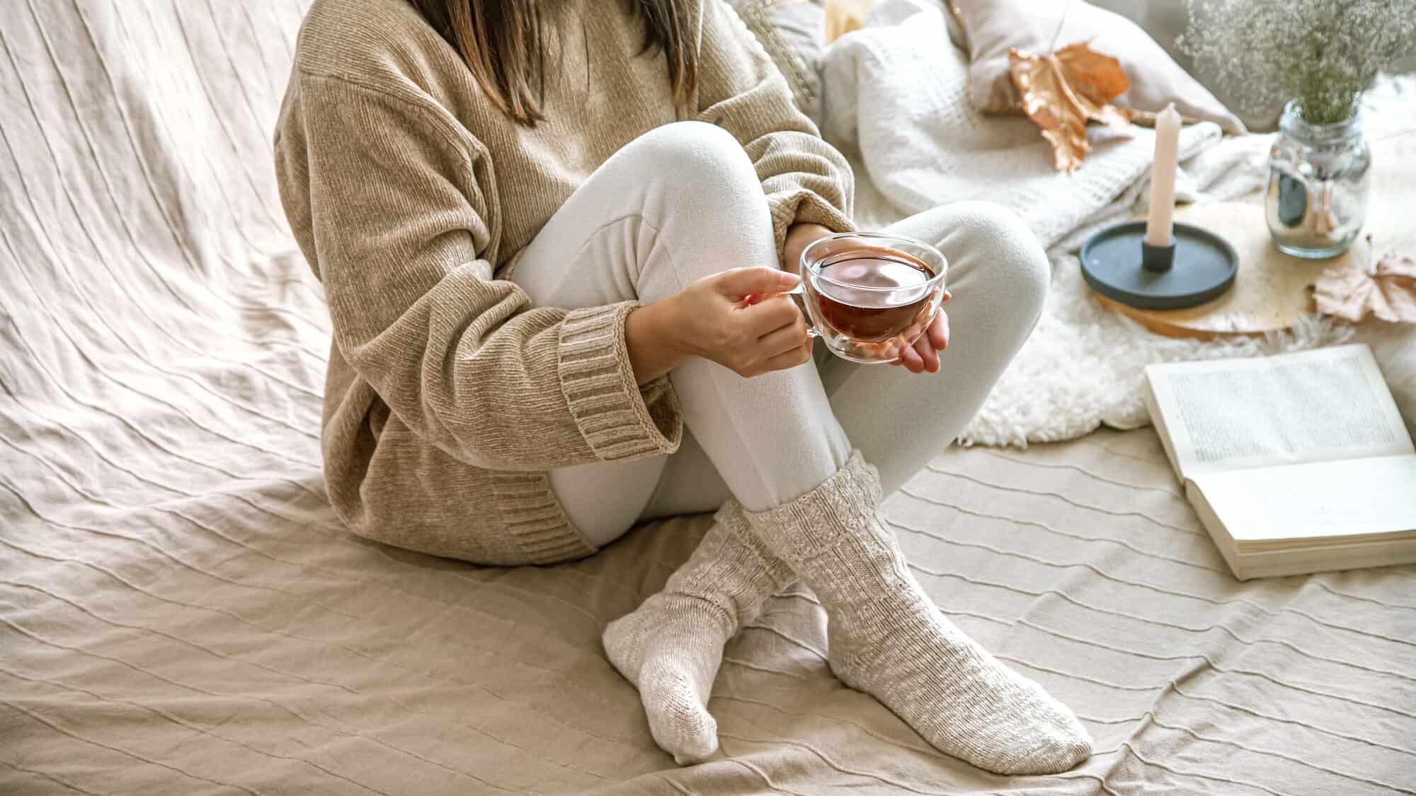 Woman in cozy sweater and socks sits on a bed holding a cup of tea, with a book and candle nearby—perfect inspiration for cozy holiday gifts.