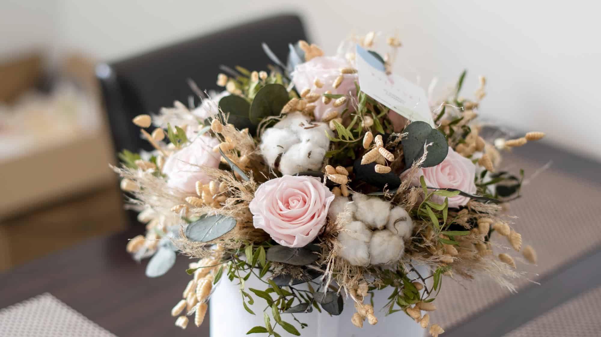 A bouquet with pink roses, cotton flowers, greenery, and dried grasses in a white vase on a table—perfect as gifts for holiday hosts and entertainers.