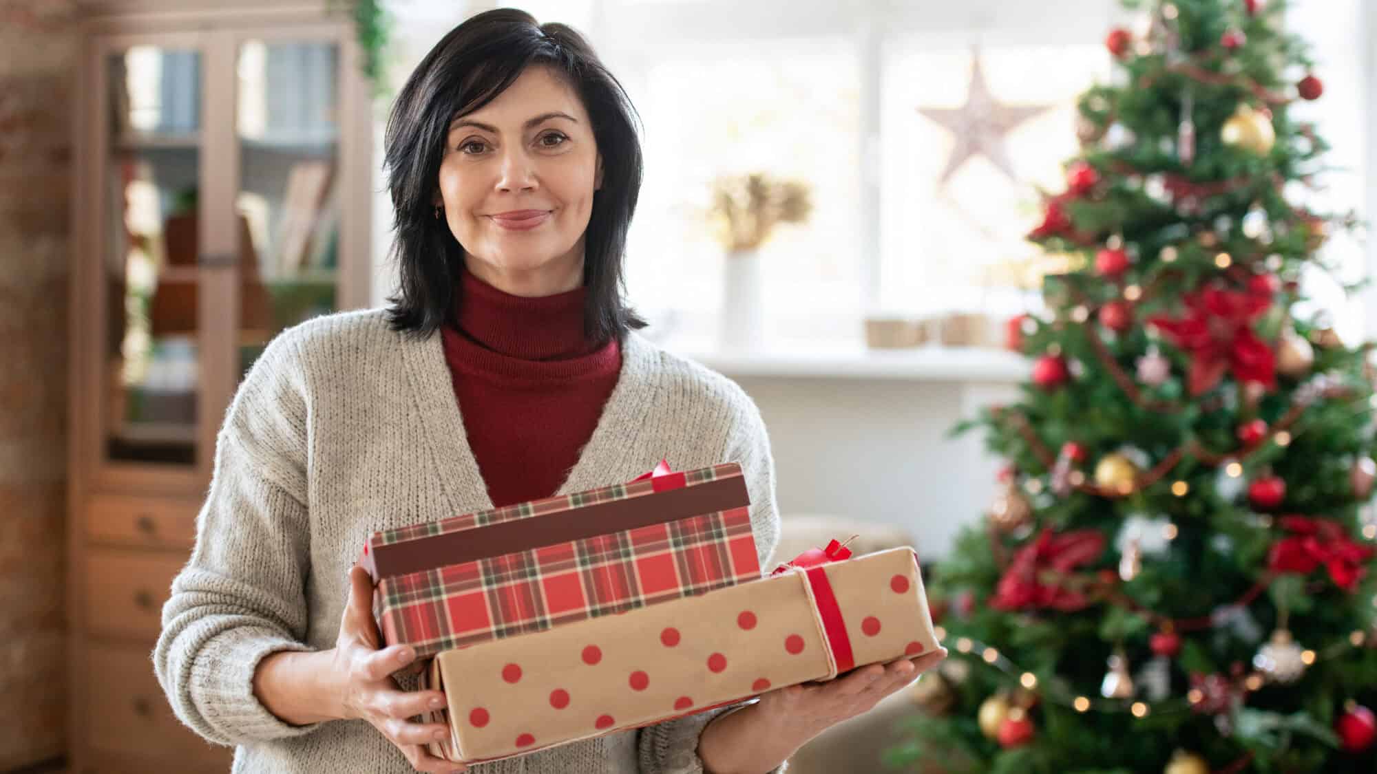 Woman holding wrapped gifts for holiday hosts and entertainers stands by a decorated Christmas tree in a cozy, festive room.