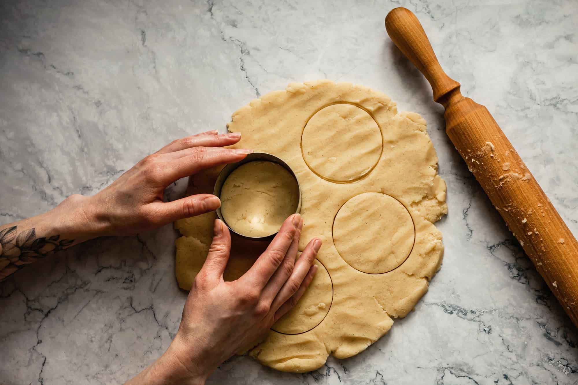 Hands cutting circles from rolled cookie dough with a round cutter&mdash;one of the essential kitchen tools for holiday baking&mdash;next to a wooden rolling pin on a marble surface.