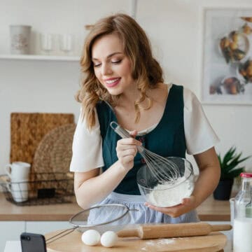 Woman whisking flour in a bowl while filming herself in a bright kitchen with baking tools and ingredients.