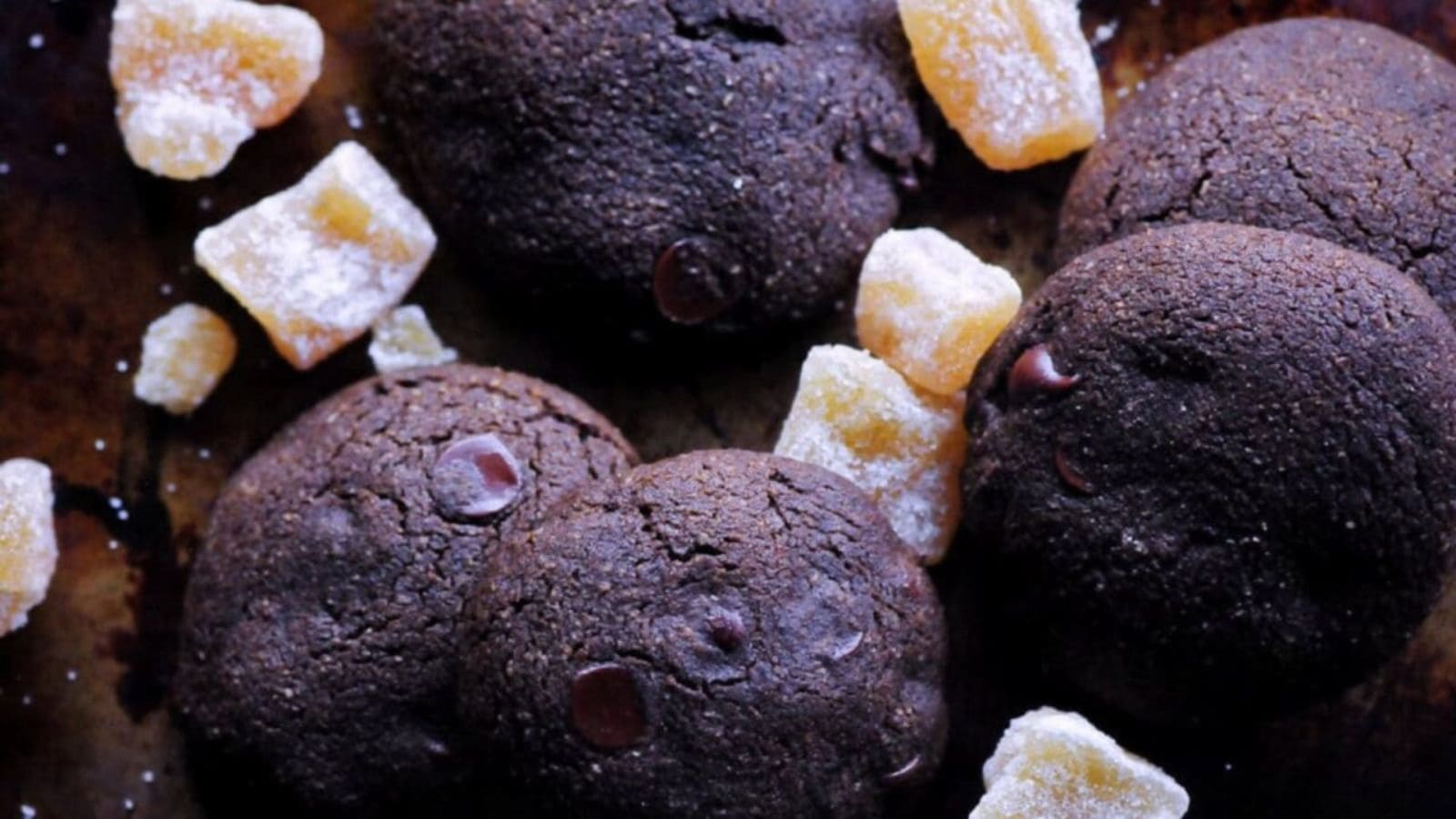 Five chocolate cookies with chocolate chips surrounded by sugar-coated ginger pieces on a dark surface.