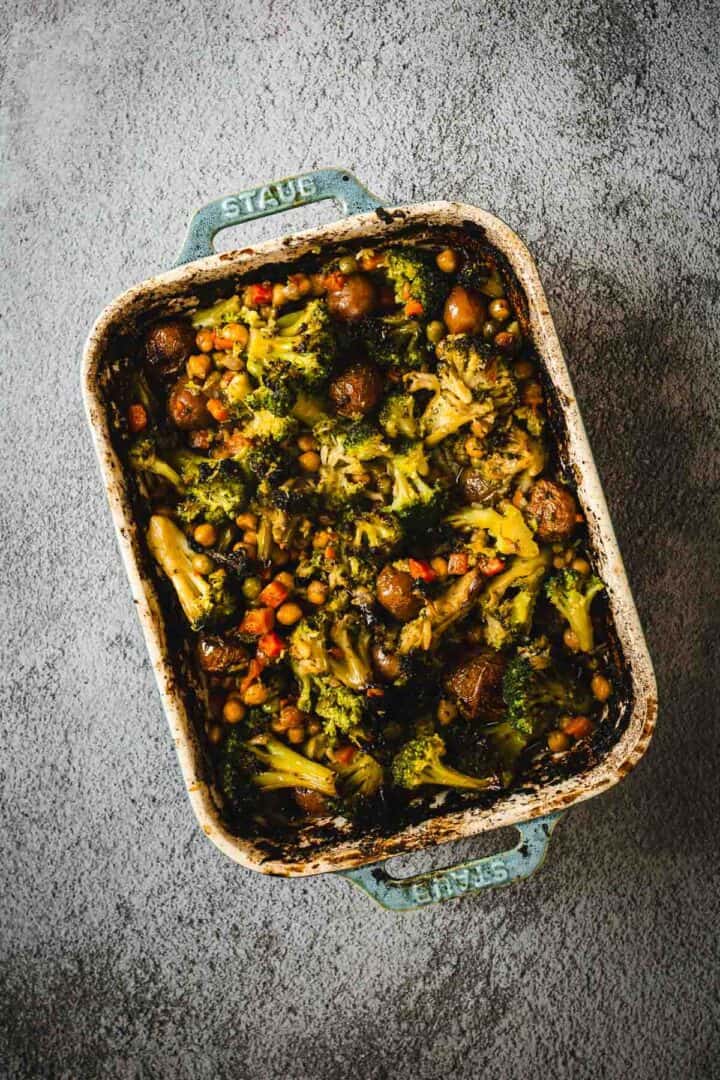 A baking dish filled with baked rice, broccoli, mushrooms, chickpeas, and Duck Fat Roasted Vegetables on a gray surface.