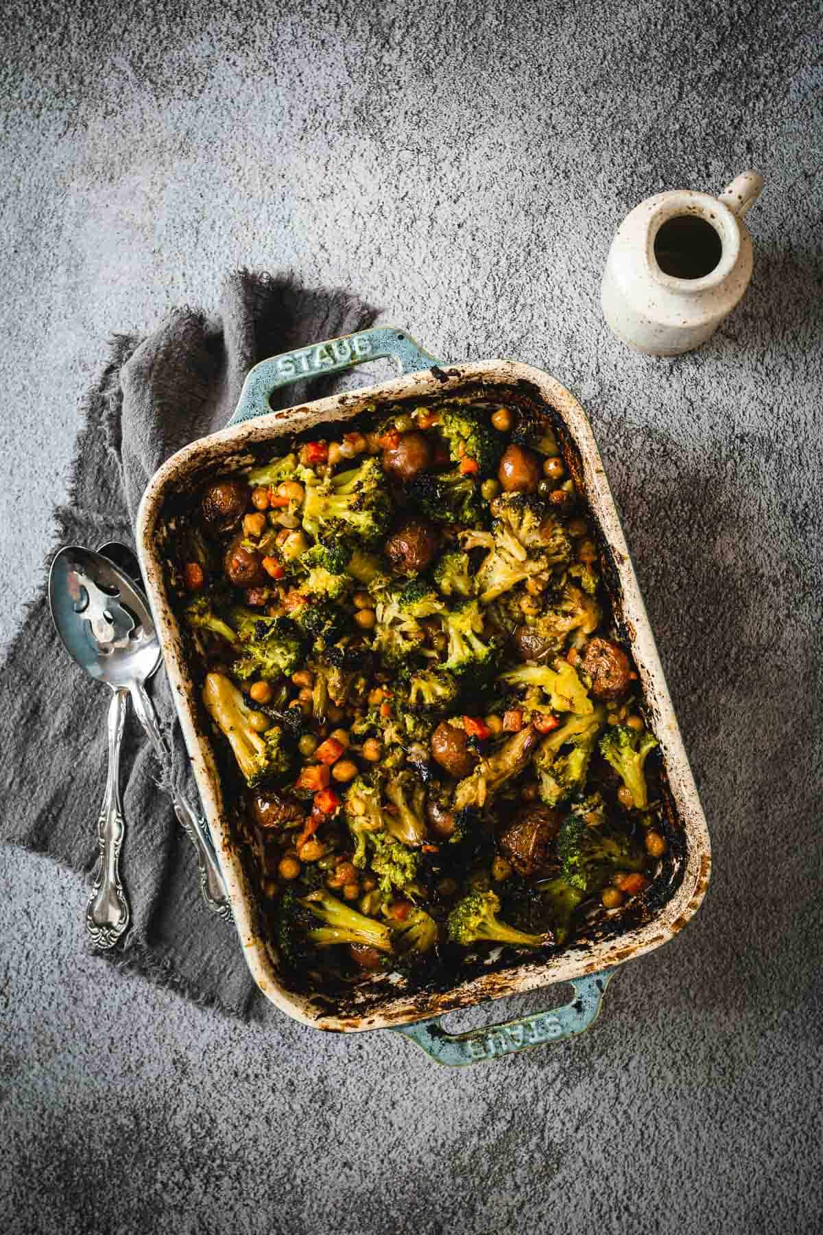Casserole dish filled with duck fat roasted vegetables and meatballs, next to serving spoons and a small pitcher.