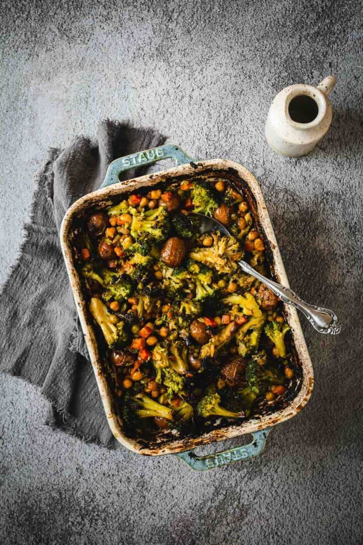 A baked vegetable casserole with broccoli, chickpeas, and mushrooms in a blue dish, reminiscent of classic Duck Fat Roasted Vegetables, with a spoon and a jug nearby.