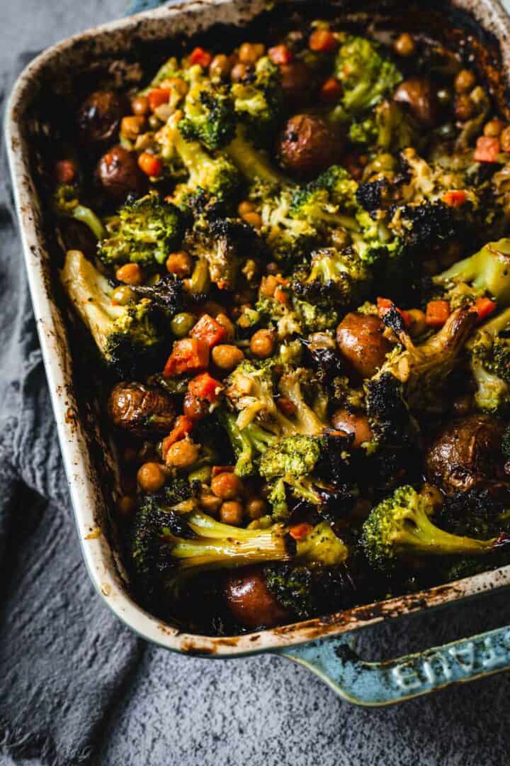 Duck Fat Roasted Vegetables, including broccoli, potatoes, chickpeas, and carrots, baked in a rustic blue casserole dish on a gray surface.
