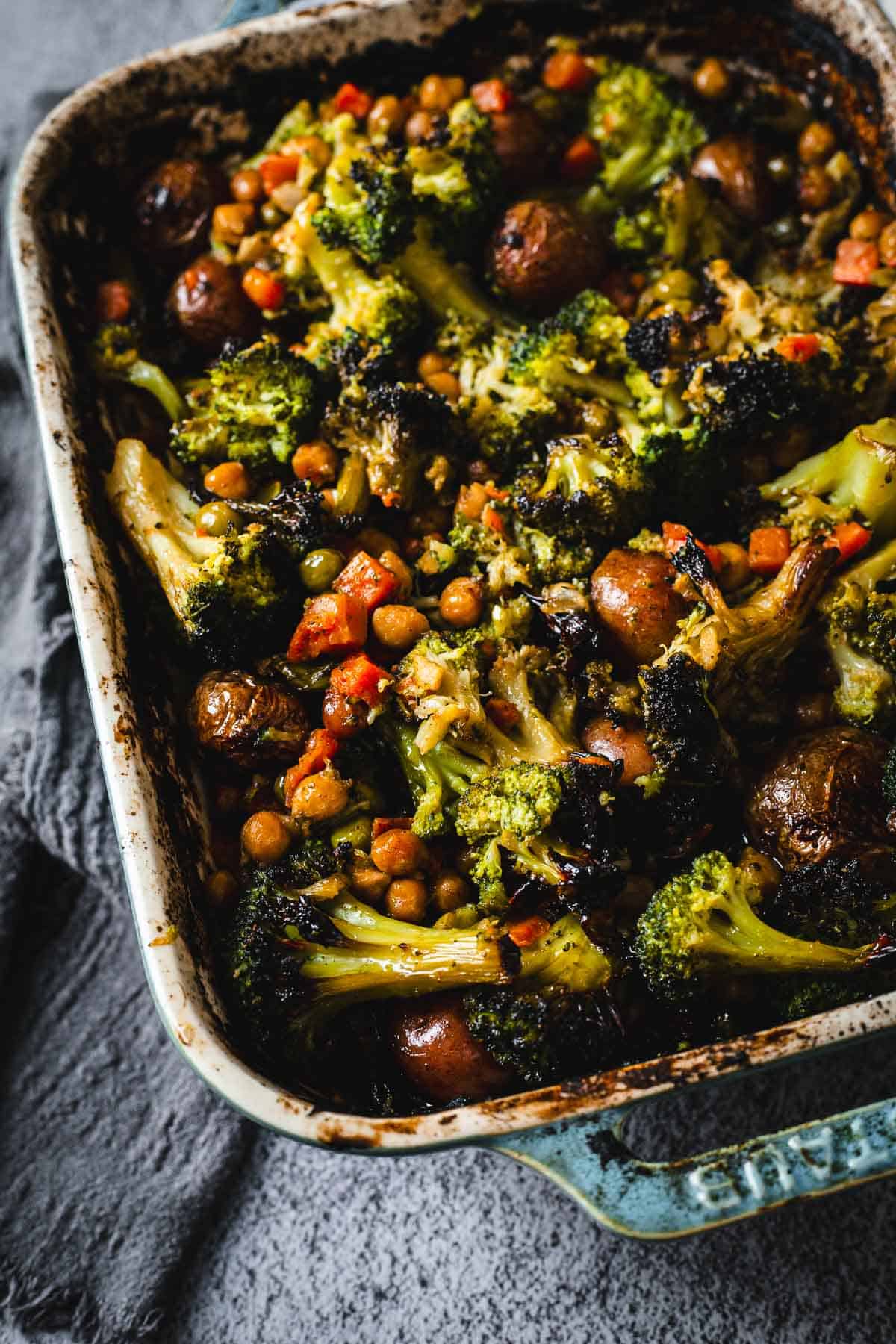 Duck Fat Roasted Vegetables, including broccoli, potatoes, chickpeas, and carrots, baked in a rustic blue casserole dish on a gray surface.