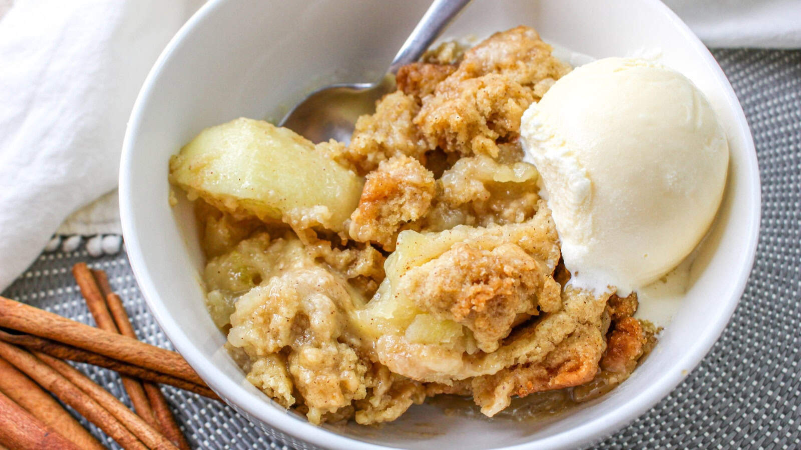 A bowl of apple crumble with a scoop of vanilla ice cream and a spoon, on a gray placemat with cinnamon sticks nearby.
