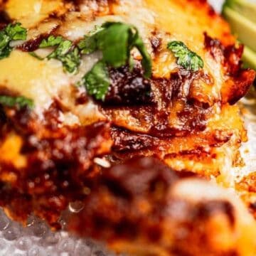 Close-up of cheesy enchiladas topped with cilantro, served with avocado slices on a clear plate.