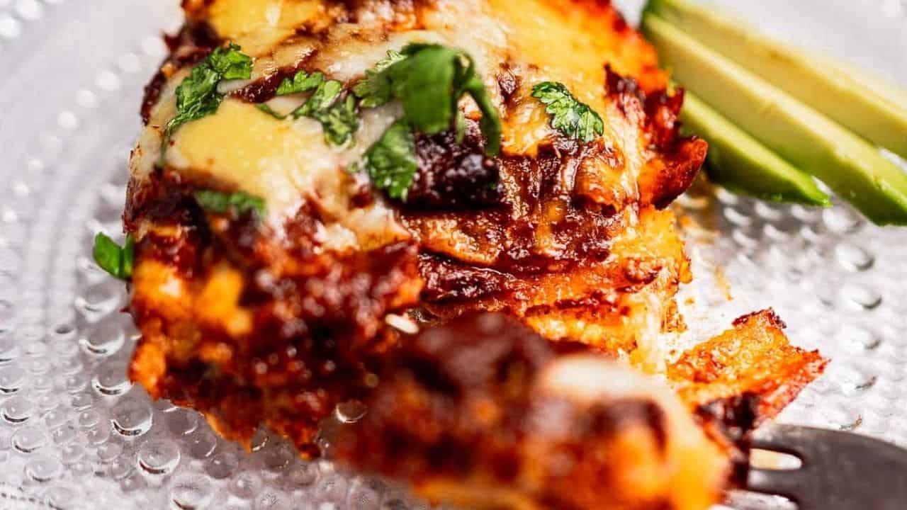 Close-up of cheesy enchiladas topped with cilantro, served with avocado slices on a clear plate.