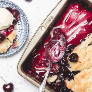 A cherry cobbler in a baking dish with a serving on a plate topped with a scoop of vanilla ice cream.