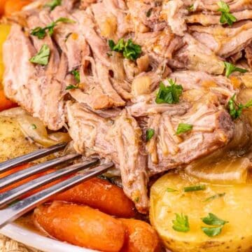 Fork beside shredded pot roast, baby carrots, and potatoes garnished with chopped parsley on a white plate.