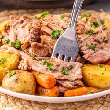 A fork shreds tender pot roast with potatoes and carrots, garnished with parsley on a white plate.