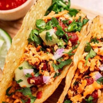 Three crunchy tacos filled with shredded veggies, sauce, and herbs, with lime wedges and salsa on the side.