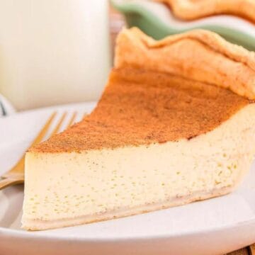A slice of custard pie with a golden crust on a white plate, beside a fork and cinnamon sticks.