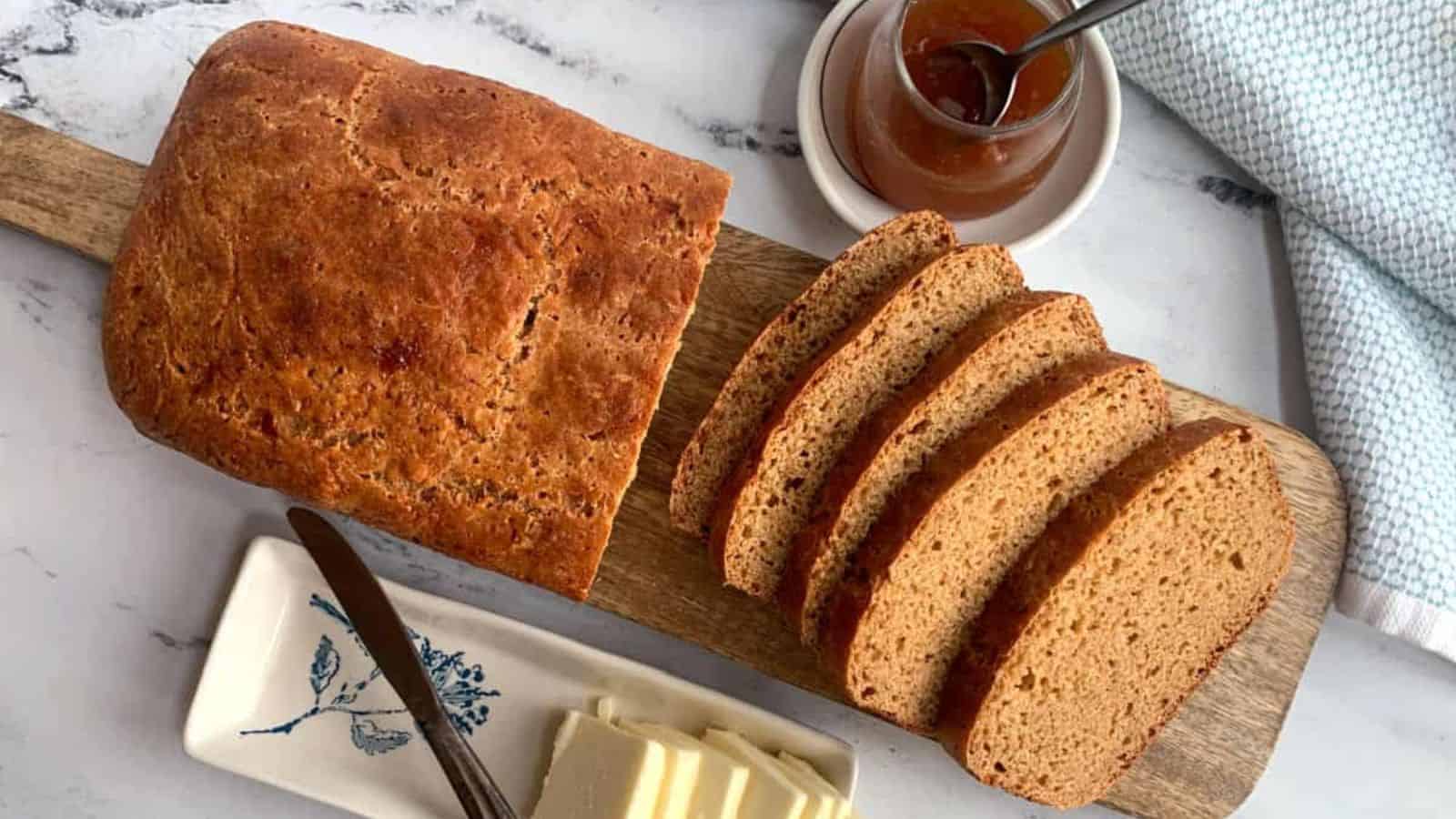 A loaf of bread, partly sliced, on a wooden board with butter and jam on the side.