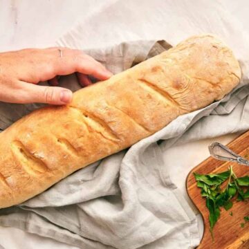 A hand reaches for a fresh baguette on a cloth, next to a bowl of herb butter and chopped parsley.
