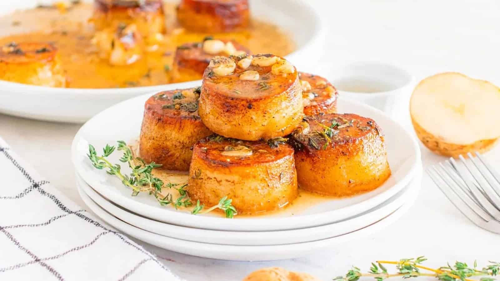 Stacked rounds of roasted potatoes garnished with herbs and garlic on a white plate, with sauce drizzled on top.