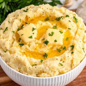 Bowl of mashed potatoes topped with melted butter and chopped parsley, with garlic bulbs in the background.