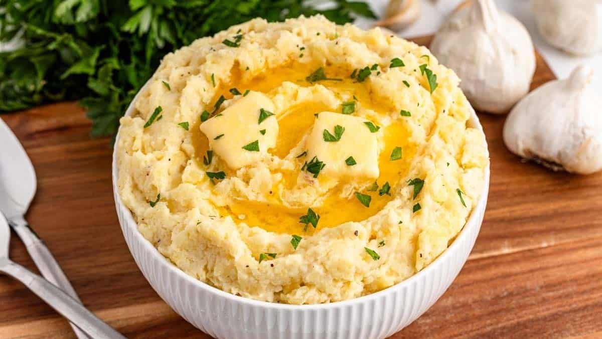 Bowl of mashed potatoes topped with melted butter and chopped parsley, with garlic bulbs in the background.