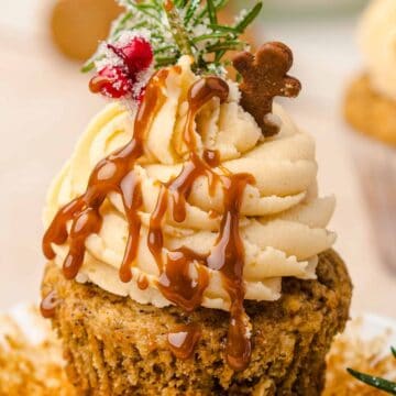 Cupcake with tan frosting, caramel drizzle, a mini gingerbread man, and festive holiday decorations on top.