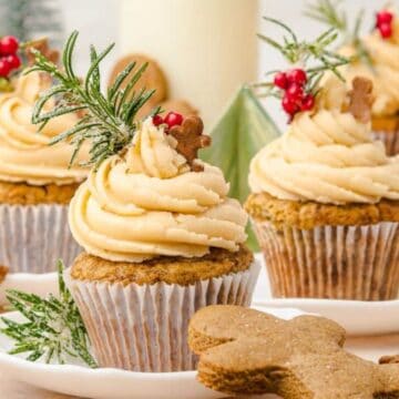 Cupcakes with cream frosting, rosemary, berries, and gingerbread cookies on white plates, festive background.
