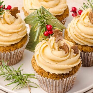 Cupcakes with creamy frosting, mini gingerbread cookies, and festive greenery on a white plate.