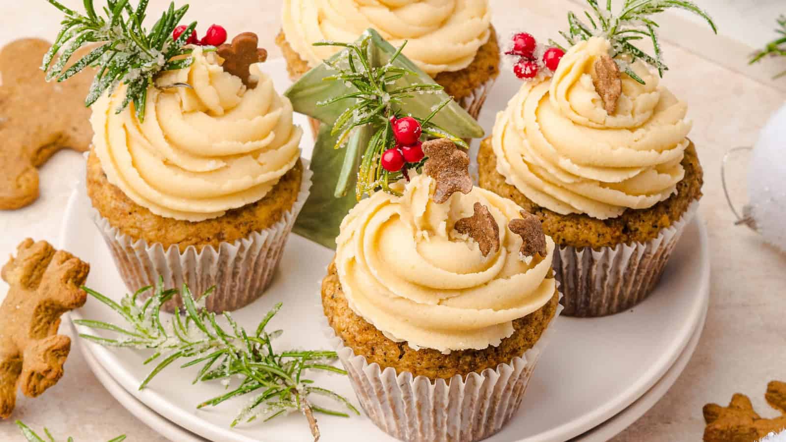 Cupcakes with creamy frosting, mini gingerbread cookies, and festive greenery on a white plate.