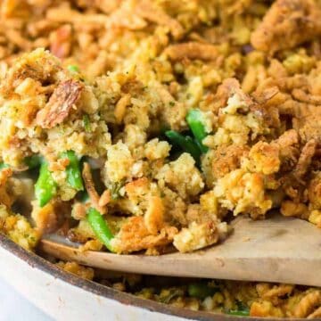 A wooden spoon scoops a serving of green bean casserole topped with crispy stuffing from a baking dish.