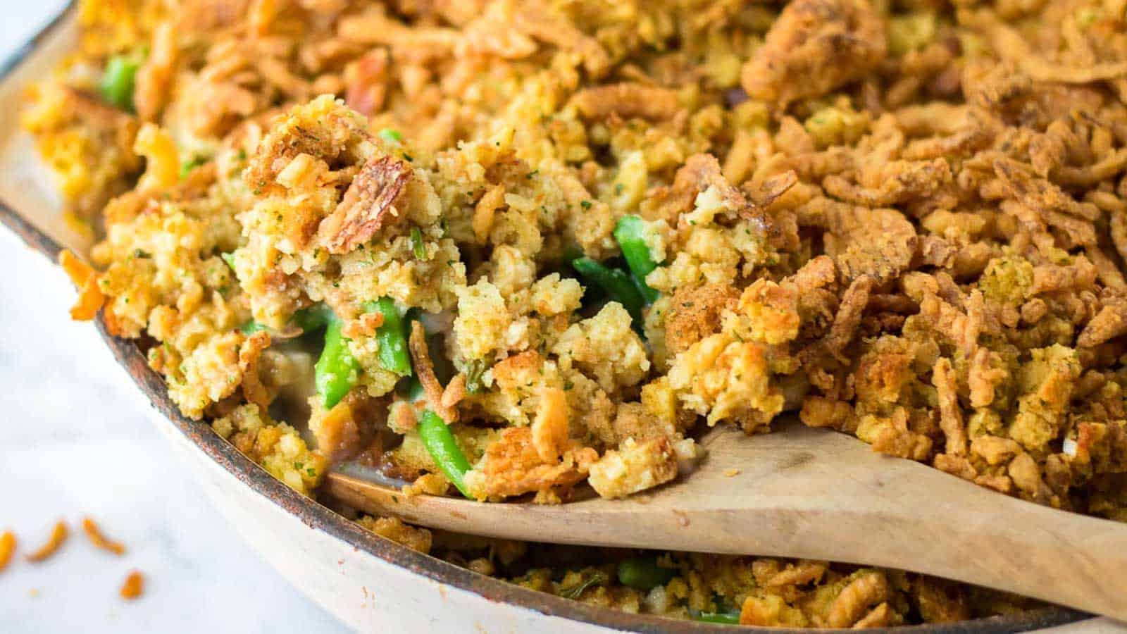 A wooden spoon scoops a serving of green bean casserole topped with crispy stuffing from a baking dish.