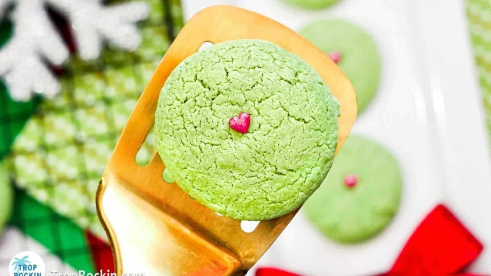 Grinch Sugar Cookies with a red candy sprinkle on top displayed on a spatula.