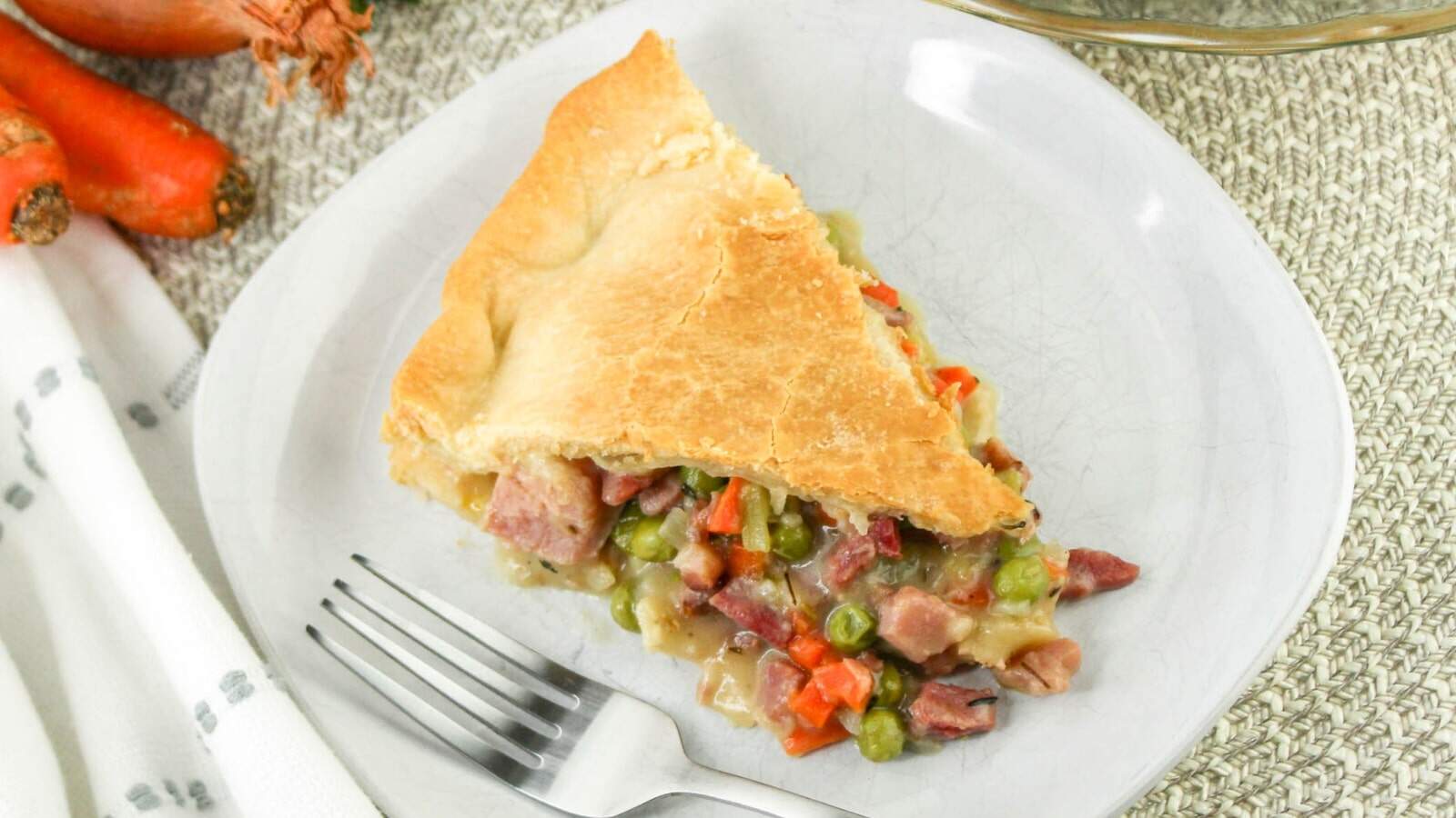 A slice of meat and vegetable pie on a plate, featuring a flaky crust and filling of diced meat, peas, and carrots. A fork is placed beside the slice.